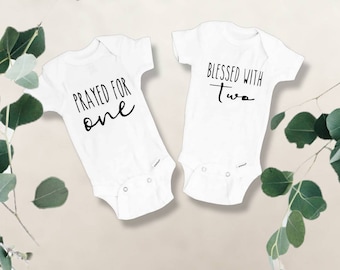 May include: Two white baby onesies with black text. One onesie says "Prayed for one" and the other says "Blessed with Two". The onesies are short-sleeved and have snap closures at the bottom. Green leaves are in the background.