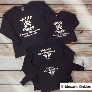 Puede incluir: Tres camisas de manga larga en azul marino con texto y gr&aacute;ficos blancos. La primera camisa tiene una huella de pata de lobo y el texto "Wolf Pack Family Weekend Wisconsin Dells". La segunda camisa tiene el mismo dise&ntilde;o pero es m&aacute;s peque&ntilde;a. La tercera camisa tiene un gr&aacute;fico de vaca y el texto "EbAcres It's a herd thing".