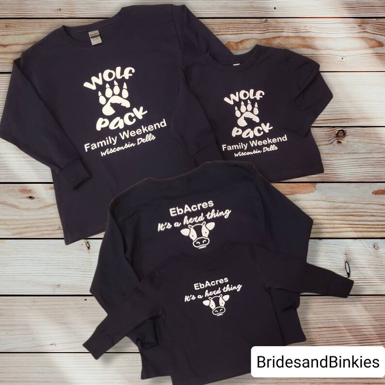 Puede incluir: Tres camisas de manga larga en azul marino con texto y gr&aacute;ficos blancos. La primera camisa tiene una huella de pata de lobo y el texto "Wolf Pack Family Weekend Wisconsin Dells". La segunda camisa tiene el mismo dise&ntilde;o pero es m&aacute;s peque&ntilde;a. La tercera camisa tiene un gr&aacute;fico de vaca y el texto "EbAcres It's a herd thing".