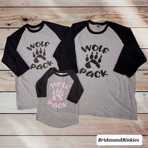 Puede incluir: Tres camisetas de b&eacute;isbol grises y negras con el texto "Wolf Pack" y un dise&ntilde;o de huella de pata. Las camisetas son de diferentes tama&ntilde;os.