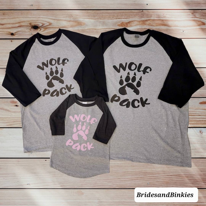 Puede incluir: Tres camisetas de b&eacute;isbol grises y negras con el texto "Wolf Pack" y un dise&ntilde;o de huella de pata. Las camisetas son de diferentes tama&ntilde;os.