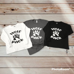 Puede incluir: Tres camisas de manga larga con el texto "Wolf Pack" y un dise&ntilde;o de huella de pata. Las camisas son blancas, grises y negras.