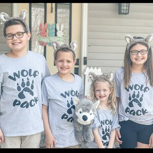 Puede incluir: Cuatro ni&ntilde;os con camisetas grises con una huella de pata de lobo y el texto "Wolf Pack" en ellas. Llevan orejas de lobo y est&aacute;n de pie frente a una casa. El ni&ntilde;o del medio sostiene un animal de peluche gris en forma de lobo.