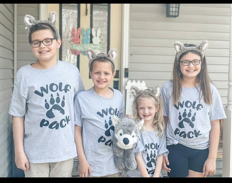 Puede incluir: Cuatro ni&ntilde;os con camisetas grises con una huella de pata de lobo y el texto "Wolf Pack" en ellas. Llevan orejas de lobo y est&aacute;n de pie frente a una casa. El ni&ntilde;o del medio sostiene un animal de peluche gris en forma de lobo.
