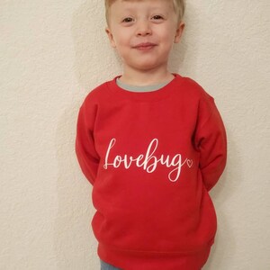 May include: A young child wearing a red sweatshirt with the text "lovebug" in white lettering and a small heart. The child is wearing blue jeans.