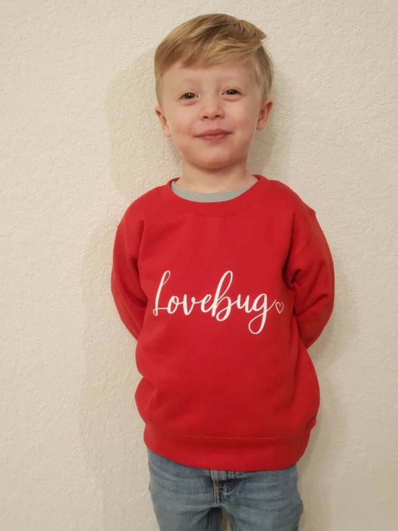 May include: A young child wearing a red sweatshirt with the text "lovebug" in white lettering and a small heart. The child is wearing blue jeans.