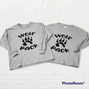 Puede incluir: Dos camisas grises de manga larga con el texto "Wolf Pack" y un gr&aacute;fico de huella de lobo negro.
