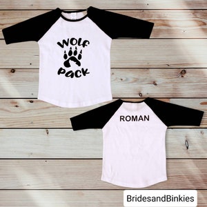Puede incluir: Dos camisetas de b&eacute;isbol blancas con mangas negras. Una camiseta tiene una huella de pata negra con el texto "Wolf Pack" en la parte delantera. La otra camiseta tiene el nombre "Roman" en la parte posterior.