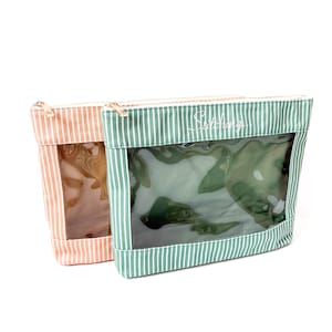 Puede incluir: Dos neceseres a rayas, uno melocotón y blanco, el otro verde y blanco. Ambos tienen una ventana transparente y cremalleras doradas. La palabra "Stitching" está escrita en la bolsa verde.