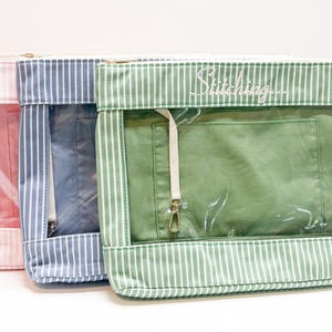 May include: Three rectangular zippered pouches in pink, blue, and green, each featuring a white vertical pinstripe pattern and a clear plastic front window. The green pouch displays the word Stitching in white cursive script above the window.