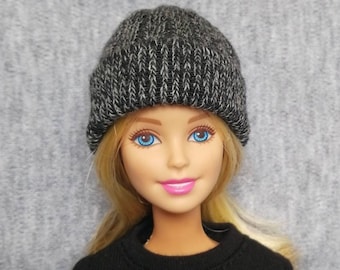Puede incluir: Una muñeca rubia con un gorro negro con un patrón de motas grises. La muñeca tiene ojos azules y labios rosados.