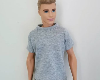 May include: A gray short-sleeved t-shirt for a doll. The t-shirt has a crew neck and is made of a soft, textured fabric.