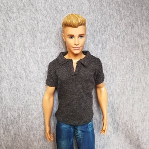 May include: A doll with short blond hair wearing a dark gray polo shirt and blue jeans.