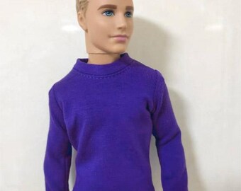 May include: A doll with blonde hair and blue eyes wearing a purple long-sleeved shirt and light blue jeans. The doll is posed against a white background. The shirt has a crew neck.