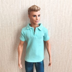 May include: A doll wearing a light blue polo shirt with a button-up collar and blue jeans.