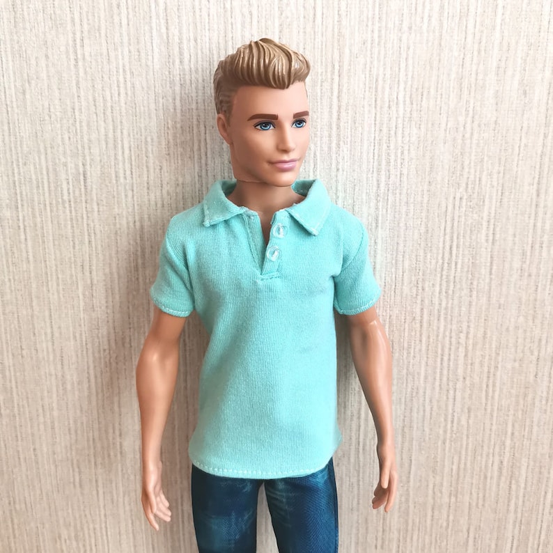 May include: A doll wearing a light blue polo shirt with a button-up collar and blue jeans.