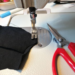 K&ouml;nnte beinhalten: Nahaufnahme einer N&auml;hmaschine mit einem schwarzen Stoff, der gen&auml;ht wird. Neben der Maschine liegt ein Paar rote Schere auf dem Tisch.