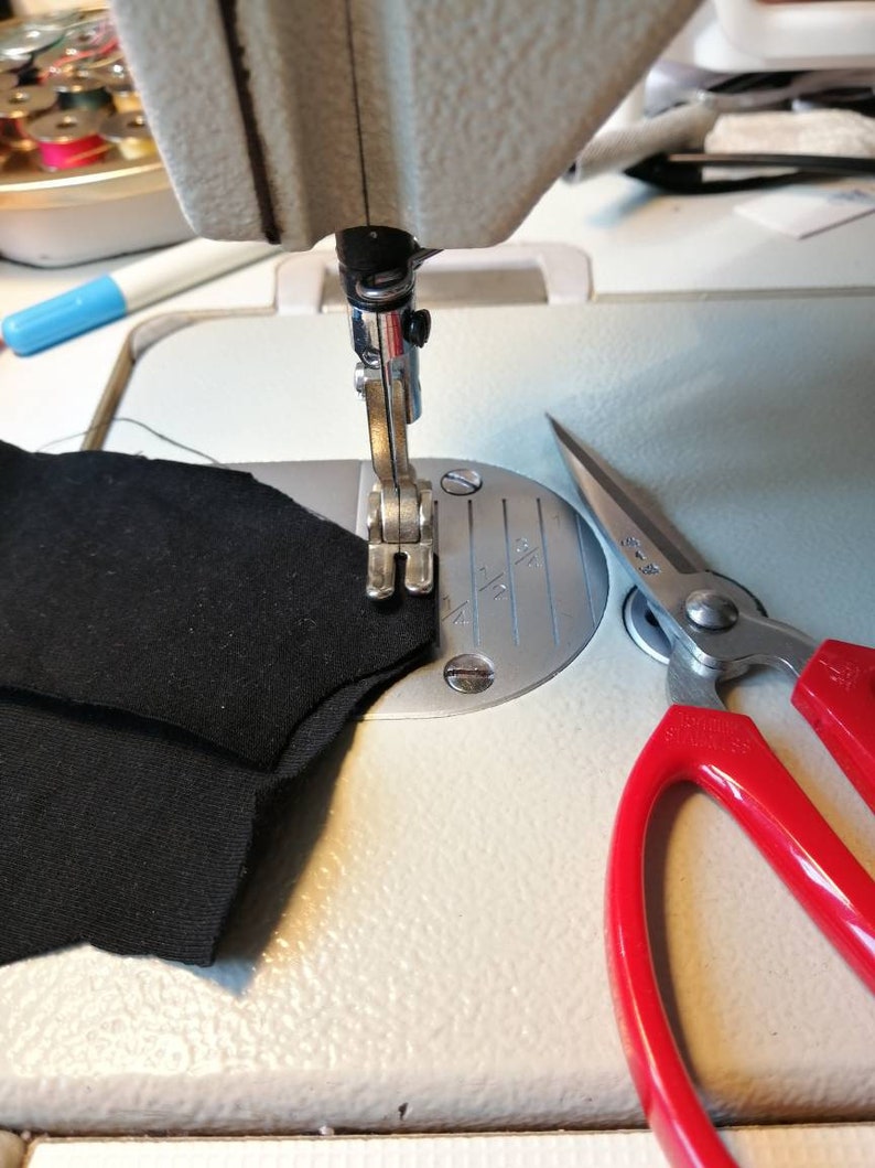 K&ouml;nnte beinhalten: Nahaufnahme einer N&auml;hmaschine mit einem schwarzen Stoff, der gen&auml;ht wird. Neben der Maschine liegt ein Paar rote Schere auf dem Tisch.