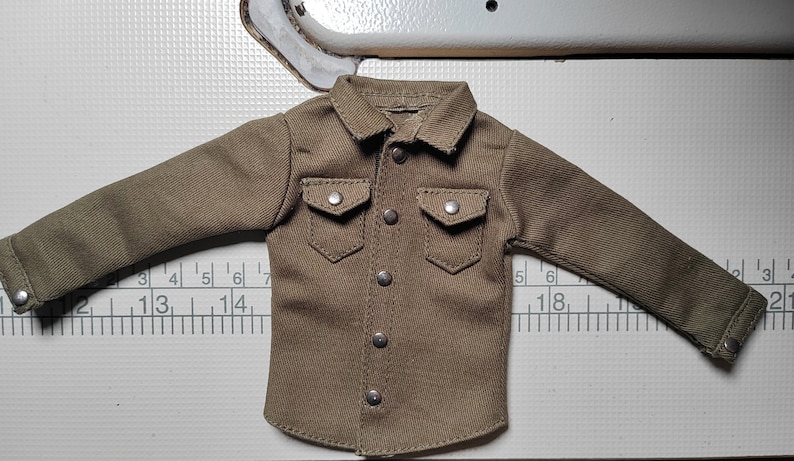 Pode incluir: Uma camisa marrom de manga comprida com fecho de bot&otilde;es e dois bolsos com aba. A camisa tem um tecido ligeiramente texturizado e &eacute; feita para uma boneca ou figura de a&ccedil;&atilde;o.