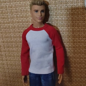 May include: A doll wearing a white and red baseball shirt with blue jeans.