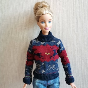 May include: A doll wearing a blue and red knitted sweater with a reindeer design and blue jeans.