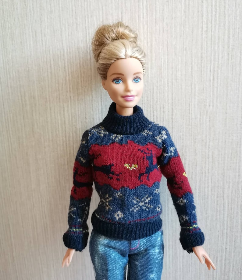 May include: A doll wearing a blue and red knitted sweater with a reindeer design and blue jeans.