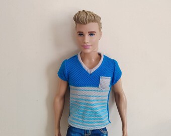May include: A doll with short blond hair wearing a blue and gray striped shirt with a blue v-neck and a small pocket on the front. The doll is wearing blue jeans.