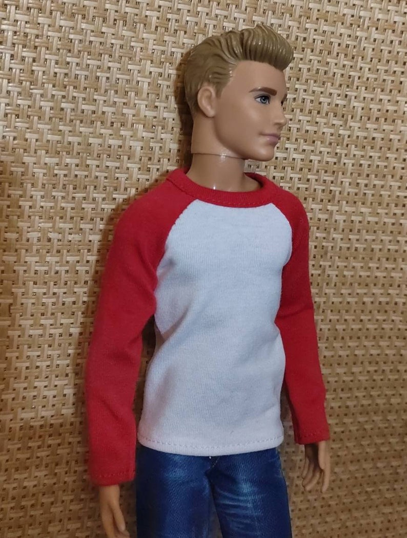 May include: A doll wearing a white long-sleeved shirt with red sleeves and blue jeans.