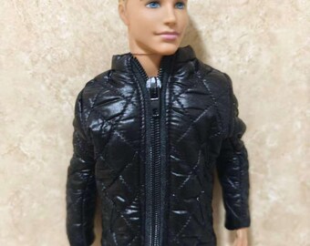May include: A doll with blonde hair and blue eyes wearing a black quilted jacket with a front zipper closure and blue pants. The jacket features a diamond-stitched pattern and a high collar. The doll has a neutral expression and light skin tone.