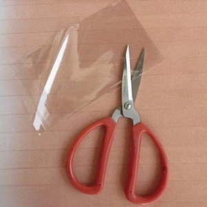 May include: Red handled scissors with silver blades, lying on a wooden surface next to a clear plastic bag.