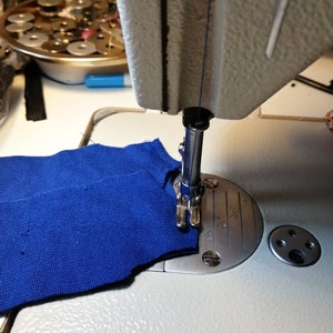 May include: A close-up of a sewing machine needle going through a piece of blue fabric. The needle is in the down position and the fabric is being held in place by the presser foot.