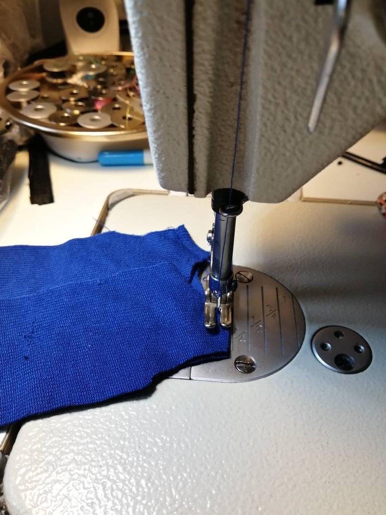May include: A close-up of a sewing machine needle going through a piece of blue fabric. The needle is in the down position and the fabric is being held in place by the presser foot.