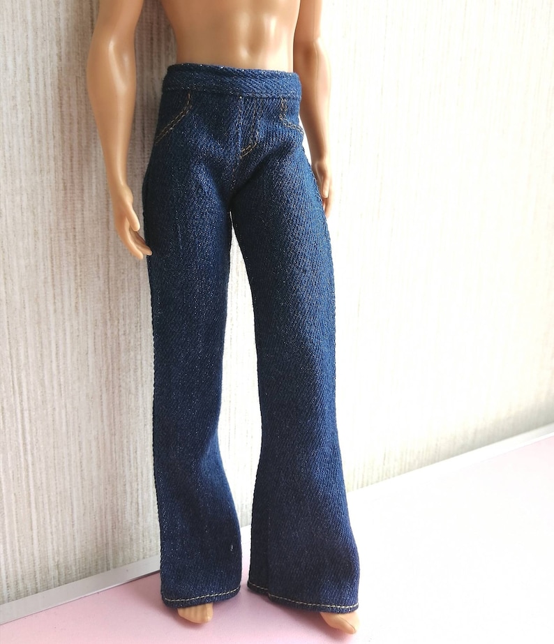 May include: Dark blue denim doll pants with a flared leg design. The pants have yellow stitching details and a classic five-pocket style. The doll pants are made of a textured denim fabric.