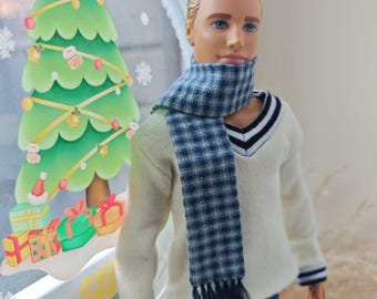 Puede incluir: Muñeco con un suéter color crema, una bufanda a cuadros azul y blanco y vaqueros azules. El muñeco tiene el pelo rubio y ojos azules. Una decoración de árbol de Navidad está en el fondo.