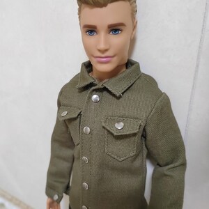 Pode incluir: Uma boneca com cabelo loiro vestindo uma camisa verde abotoada e cal&ccedil;as pretas.