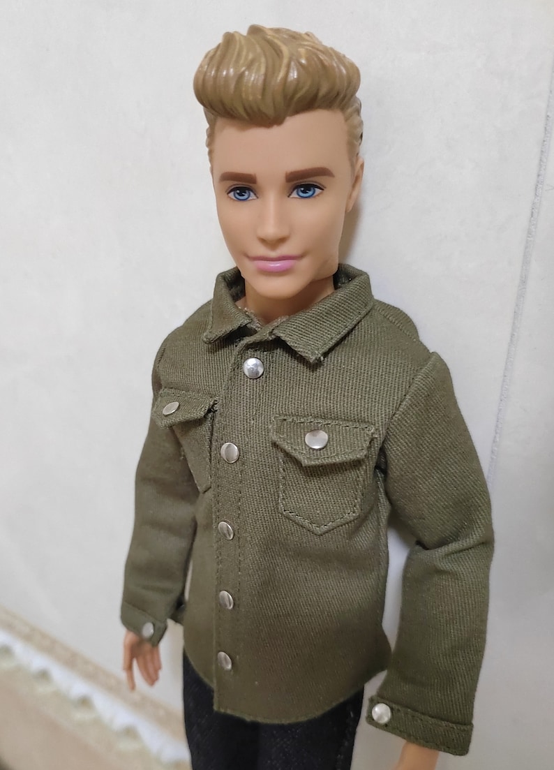 Pode incluir: Uma boneca com cabelo loiro vestindo uma camisa verde abotoada e cal&ccedil;as pretas.