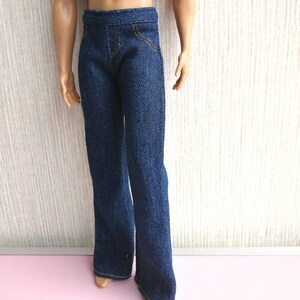 May include: Dark blue denim pants with a straight leg cut. The pants have a classic five-pocket design with gold stitching. The denim fabric appears to be a medium weight. The pants are shown on a doll.