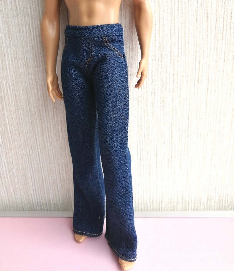 May include: Dark blue denim pants with a straight leg cut. The pants have a classic five-pocket design with gold stitching. The denim fabric appears to be a medium weight. The pants are shown on a doll.