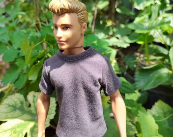 May include: A doll wearing a gray t-shirt and blue jeans. The doll has short, blond hair and blue eyes.