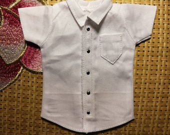 Pode incluir: Uma camisa branca de manga curta com botões e um bolso no lado esquerdo. A camisa tem colarinho e seis botões pretos na frente.