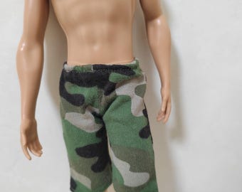 Puede incluir: Pantalones cortos de camuflaje para una muñeca. Los pantalones cortos son verdes, negros y grises.