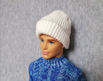 Puede incluir: Un gorro blanco de punto con textura de canalé. El gorro está puesto en una muñeca con ojos azules y un suéter azul.