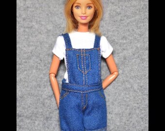 Può includere: Una bambola bionda con una maglietta bianca e una salopette di jeans blu. La salopette ha un dettaglio di cucitura marrone.