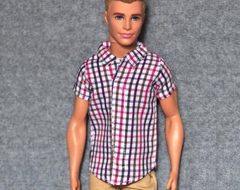 May include: A doll wearing a short-sleeved plaid shirt with red, brown, and white squares. The shirt has a button-up front and a collar. The doll is also wearing khaki pants.