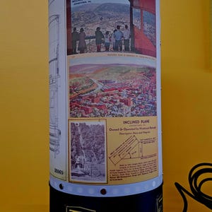 May include: Cylindrical lamp with images and text related to the Inclined Plane in Johnstown, PA. The lamp features vintage photographs and diagrams, with the text "INCLINED PLANE JOHNSTOWN PA" and "BETHLEHEM STEEL". The lamp has a black base and a white top.