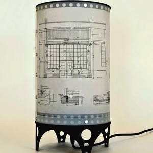 May include: A table lamp with a cylindrical shade featuring a blueprint design. The shade has a silver-grey finish with a black base and a black cord.