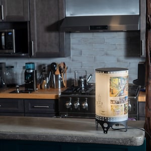 May include: A lamp with a cylindrical shade featuring a vintage postcard design of a city street scene. The lamp is on a counter in a kitchen setting.