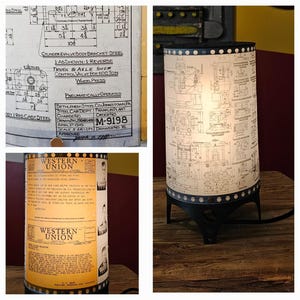 May include: A table lamp with a white lampshade featuring a blueprint design. The lampshade is surrounded by a black band with silver studs. The lamp base is black metal with three legs.