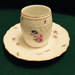 May include: A white porcelain cup and saucer set with a floral design. The cup has a gold rim and the saucer has a gold edge.