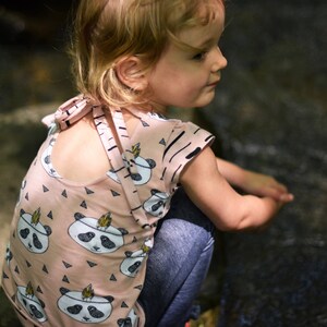 May include: A young girl wearing a pink and black panda print shirt with a criss-cross back detail. The shirt has a short sleeve and a relaxed fit.